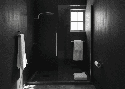 Modern black bathroom featuring a frameless glass shower, matte textured walls, and a polished stone floor with delicate droplets. Natural light enters through a small window, illuminating soft towels, creating a serene and inviting ambiance.
