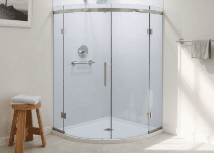 Frameless glass shower in softly lit bathroom with stone-tiled floor, brushed metal fixtures, and white tile accents, complemented by a wooden stool, creating a warm and inviting relaxation space.